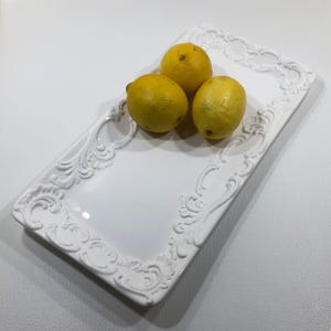 May include: A rectangular white ceramic serving tray featuring an intricate raised scrollwork border design along the edges. Three bright yellow lemons are positioned on the smooth, glossy surface of the tray.