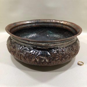 May include: A copper-toned bowl with intricate embossed elephant and floral designs. The bowl has a wide rim and a dark interior. A small gold ring with three stones sits beside the bowl.