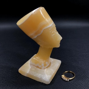 May include: A small, yellow alabaster sculpture of an Egyptian queen's head, wearing a traditional headdress. The sculpture is on a white alabaster base.