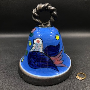 May include: A blue ceramic bell with a black twisted handle. The bell is decorated with a colorful bird design.