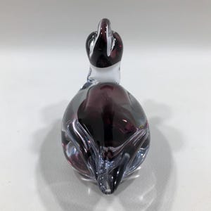 May include: A decorative glass duck figurine. The duck is primarily purple with clear and white accents. The glass is shaped to resemble a duck with a long neck and a rounded body. The figurine is on a white surface.