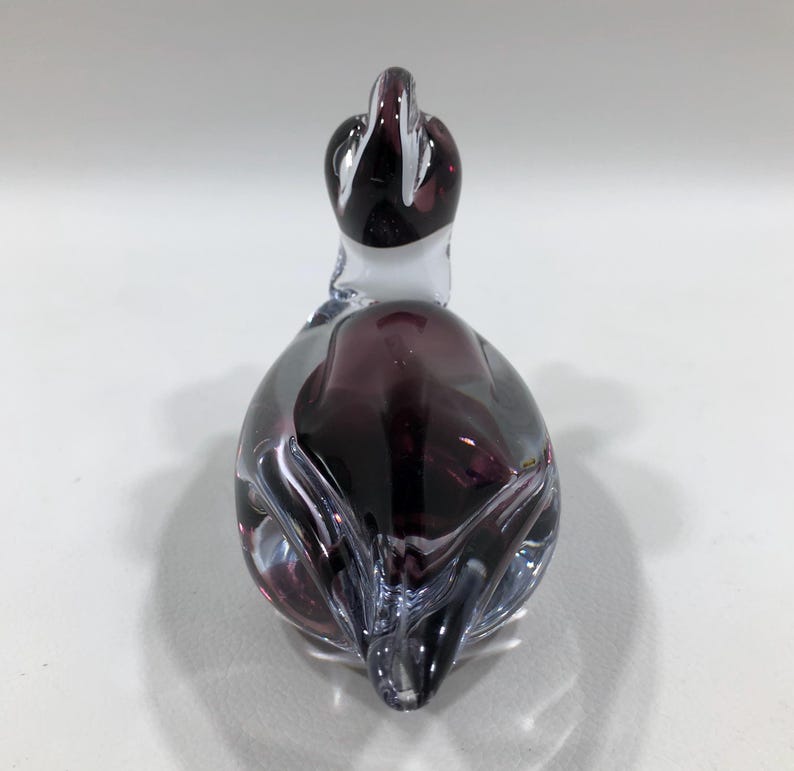 May include: A decorative glass duck figurine. The duck is primarily purple with clear and white accents. The glass is shaped to resemble a duck with a long neck and a rounded body. The figurine is on a white surface.