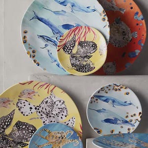 Może przedstawiać: Kolekcja okrągłych ceramicznych talerzy z akwarelowymi ilustracjami życia morskiego, w tym płetwali błękitnych, nakrapianych ryb rozdymek i ośmiornicy, na żywych ż&oacute;łtych, pomarańczowych i jasnoniebieskich tłach z koralowymi akcentami.