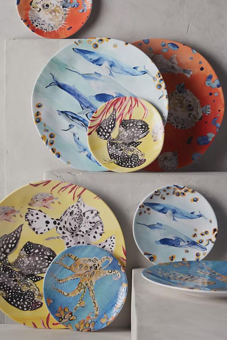 Może przedstawiać: Kolekcja okrągłych ceramicznych talerzy z akwarelowymi ilustracjami życia morskiego, w tym płetwali błękitnych, nakrapianych ryb rozdymek i ośmiornicy, na żywych ż&oacute;łtych, pomarańczowych i jasnoniebieskich tłach z koralowymi akcentami.