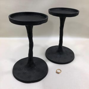 May include: Two black metal candle holders with circular bases and textured, uneven stems that resemble tree branches. Each holder features a flat, round top platform for candles. A small gold-toned ring is placed on the surface nearby.