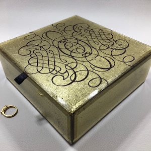 May include: A gold mirrored jewelry box with a black ornate design on the lid. The box has a black pull tab on the front.