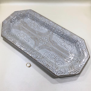 May include: An elongated octagonal ceramic tray featuring a light gray and white glaze with a repeating embossed pattern of floral motifs and geometric lines. A small gold-toned ring is positioned near the bottom left corner of the tray.