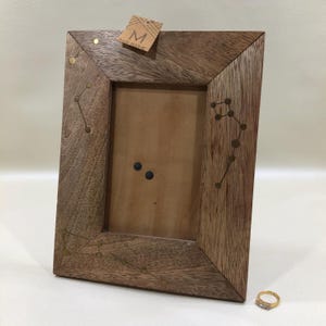 May include: Wooden picture frame with constellation designs and brass accents. The frame has a natural wood finish and a rectangular shape. A small tag with the letter "M" is attached to the top. A gold ring with small stones is in the bottom right corner.