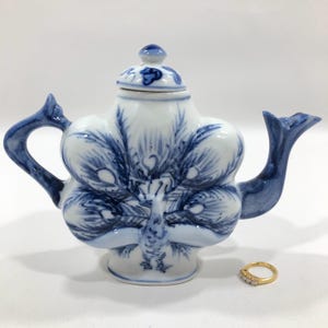 May include: A small blue and white porcelain teapot shaped like a peacock with a lid. The teapot has a blue and white design of a peacock with its tail feathers spread out.