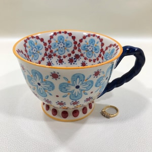 May include: A white ceramic teacup with a blue floral pattern and a dark blue handle. The rim and base are accented with orange and red details. A gold ring with small stones sits next to the cup.