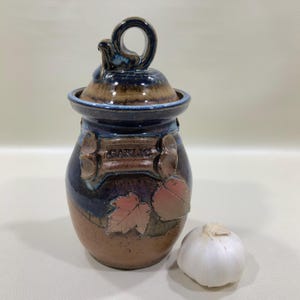 May include: A ceramic garlic keeper with a lid and handle. The jar is brown, blue, and tan with the word "GARLIC" on the front. Two leaf-shaped embellishments are on the front. A whole garlic bulb sits next to the jar.