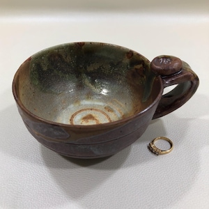 May include: Handmade ceramic mug with a brown and green glaze. The mug has a rounded shape with a handle and a decorative ring. A gold ring with small stones is next to the mug.