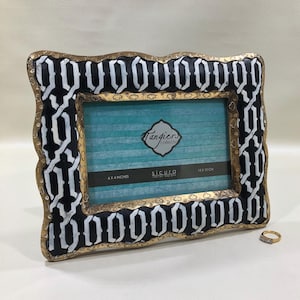 May include: Ornate picture frame with a black and white geometric pattern and gold trim. The frame holds a blue and white striped background. The frame is labeled "Tangiers collection" and is sized at 6 x 4 inches and 15 x 10 cm.