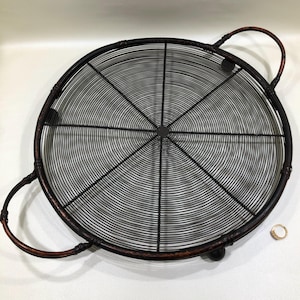 May include: A round, black metal serving tray with a wire mesh design. The tray has two curved handles and a dark brown rim. A small gold ring with a gemstone is visible in the corner.