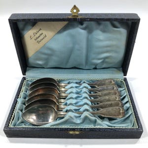 May include: A set of silver spoons in a blue velvet-lined box. The box is dark blue with a decorative clasp and a label that reads "L. Lülwes Hofjuwelier Detmold". The spoons are arranged neatly inside the box.