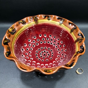 May include: A red ceramic colander with a scalloped rim and a brown glaze. The colander has a pattern of small holes in the bottom.