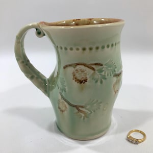 May include: A handmade ceramic mug with a light green glaze. The mug features a handle and is decorated with embossed pinecones and needles. The rim has a brown glaze, and the handle has a textured design. A gold ring with three small diamonds is next to the mug.