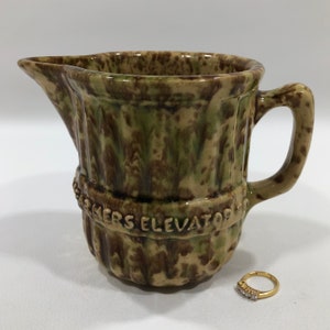 May include: A brown and green glazed ceramic pitcher with a handle. The pitcher has a band around the middle with the words "Smyers Elevator Co."