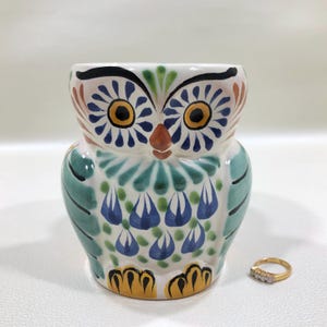 May include: A ceramic owl-shaped planter with a colorful hand-painted design. The owl features large yellow eyes, blue and green feather details, and a white base. A gold ring with three small diamonds is next to the planter.