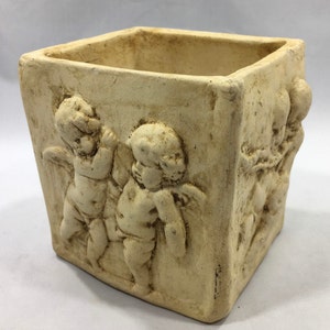May include: A square ceramic planter with a beige finish features three cherub figures in relief. The planter is ideal for displaying small plants or flowers.