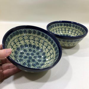 May include: Two ceramic bowls with a blue rim and a floral pattern in shades of blue and green. The bowls have a light beige background and are likely handmade. The bowls are suitable for serving food.