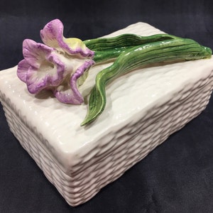 May include: A white ceramic box with a raised, woven pattern. A three-dimensional purple and yellow flower with green leaves is attached to the top of the box.