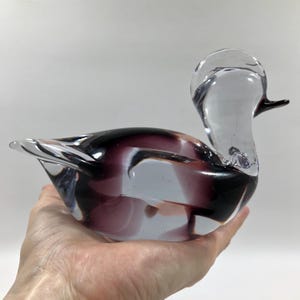 May include: A clear glass duck figurine with a purple and black interior. The duck has a long neck and a small, black beak. The figurine is held in a hand.