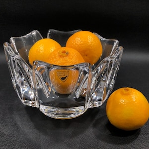 May include: A clear, faceted glass bowl with a starburst design, filled with four bright orange tangerines. The bowl has a label that reads "Orrefors Sweden". One tangerine sits outside the bowl on a black surface.