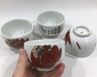 May include: Four white ceramic tea cups with red and gold accents. One cup is held by a hand. The cups feature a red and gold design and Japanese characters.
