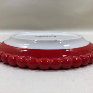 May include: A small, decorative plate with a red, beaded edge and a white interior. The plate has a glossy finish and a slightly raised rim. The red section is a vibrant, solid colour, and the plate appears to be made of ceramic.