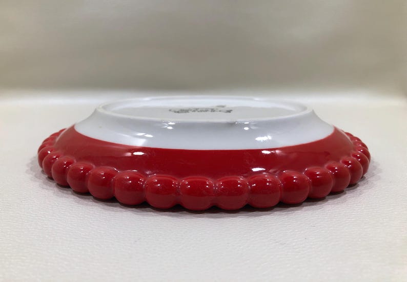 May include: A small, decorative plate with a red, beaded edge and a white interior. The plate has a glossy finish and a slightly raised rim. The red section is a vibrant, solid colour, and the plate appears to be made of ceramic.
