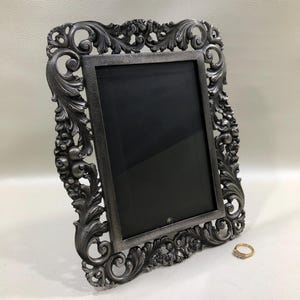 May include: A rectangular picture frame with a dark grey metallic finish and an ornate, raised scrollwork border featuring floral and leaf motifs. The frame surrounds a solid black rectangular center. A small gold ring sits near the bottom right.