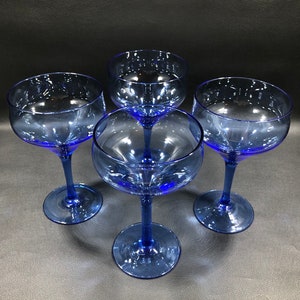 May include: Four cobalt blue glass cocktail glasses with a wide bowl and a long stem. The glasses are arranged in a circle on a black surface.
