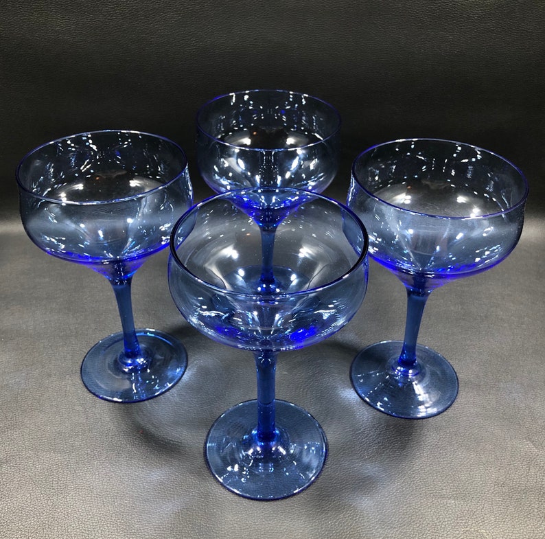 May include: Four cobalt blue glass cocktail glasses with a wide bowl and a long stem. The glasses are arranged in a circle on a black surface.
