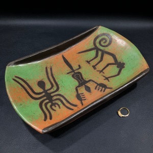 May include: A rectangular ceramic serving tray with a green and orange glaze. The tray features black painted designs of a spider, a figure holding a sword, and a stylized figure with a spiral on its head.