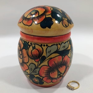 May include: A wooden trinket box with a round lid, painted with a floral design in shades of orange, red, green, and blue. The box is decorated with a red band around the top and a black and white dotted band around the bottom.