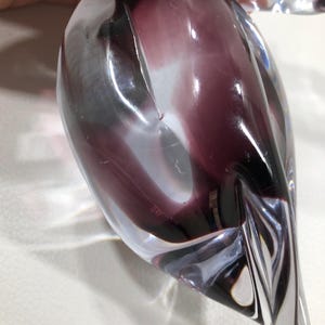 May include: A clear glass sculpture with a teardrop shape, featuring swirling shades of purple and white. The sculpture has a smooth, polished surface and is held by a hand. The design is abstract and artistic.