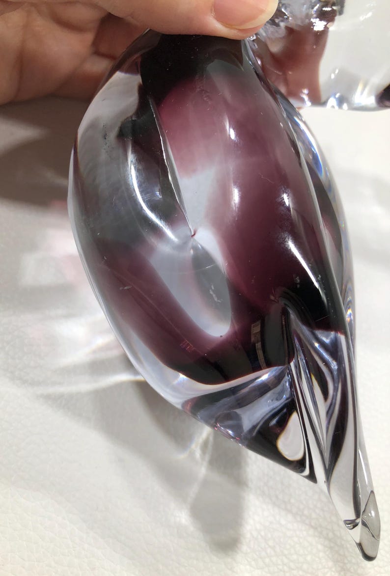 May include: A clear glass sculpture with a teardrop shape, featuring swirling shades of purple and white. The sculpture has a smooth, polished surface and is held by a hand. The design is abstract and artistic.
