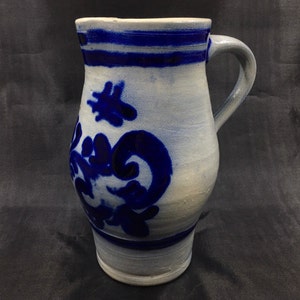 May include: A white ceramic pitcher with a blue floral design and a blue stripe around the top. The pitcher has a handle and a rounded body.