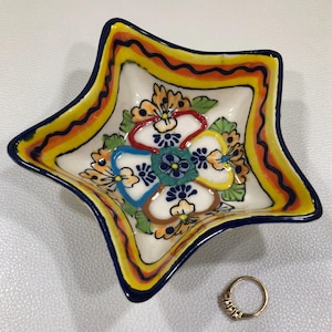 May include: A star-shaped ceramic bowl featuring a hand-painted floral design in shades of yellow, orange, blue, green, and brown, with a dark blue rim. A gold-toned ring with small embedded stones is positioned next to the bowl for scale.