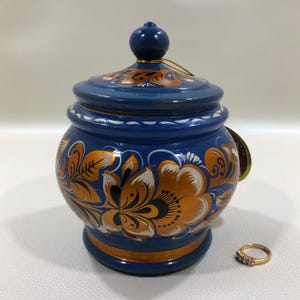 May include: A blue, lidded jar with a round knob on top. The jar is decorated with orange and white floral patterns. A gold ring with three small stones sits next to the jar.