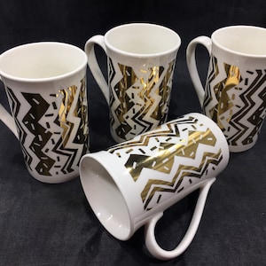 May include: Four white ceramic mugs with gold zigzag and dash patterns. The mugs have a tall, cylindrical shape with a curved handle. One mug is lying on its side, while the others stand upright, showcasing the gold design.
