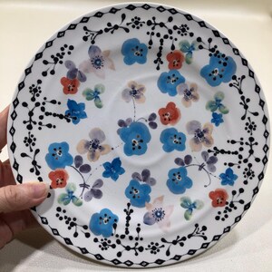 May include: White ceramic plate with a floral design. The plate features blue, red, and purple flowers, with a black decorative border. The flowers are hand-painted, adding a unique touch to the plate's design. A charming addition to any kitchen or dining room.