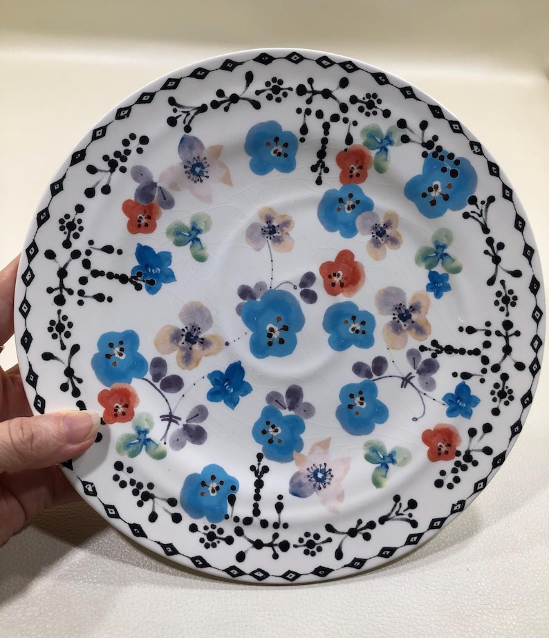 May include: White ceramic plate with a floral design. The plate features blue, red, and purple flowers, with a black decorative border. The flowers are hand-painted, adding a unique touch to the plate's design. A charming addition to any kitchen or dining room.