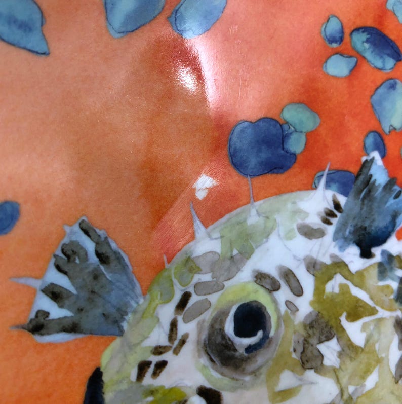 Może przedstawiać: Ceramiczny talerz z ilustracją w stylu akwareli przedstawiającą rozdymkę z zielonymi i brązowymi wzorami. Ryba znajduje się na żywym pomarańczowym tle ozdobionym rozproszonymi niebieskimi okręgami przypominającymi pęcherzyki powietrza.