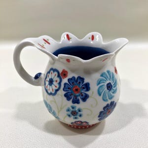 Puede incluir: Jarra de cer&aacute;mica blanca con borde festoneado y asa. La jarra est&aacute; decorada con motivos florales azules y rojos y tiene un interior azul oscuro. La base tiene un dise&ntilde;o de puntos rojos y blancos.