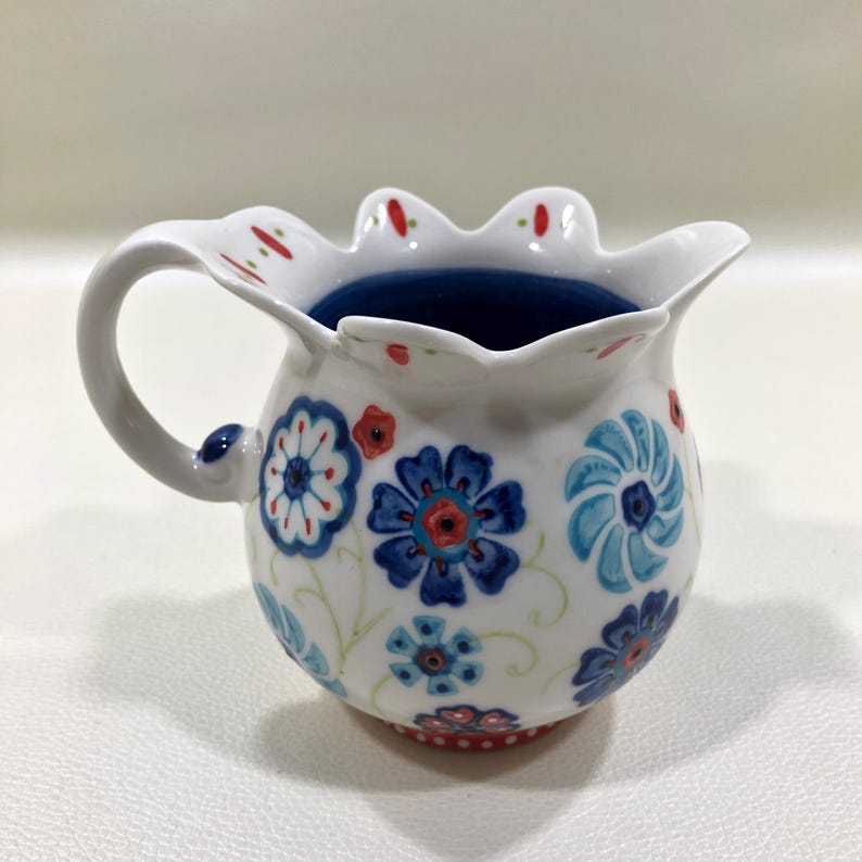 Puede incluir: Jarra de cer&aacute;mica blanca con borde festoneado y asa. La jarra est&aacute; decorada con motivos florales azules y rojos y tiene un interior azul oscuro. La base tiene un dise&ntilde;o de puntos rojos y blancos.