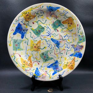 May include: A white ceramic plate with a whimsical cat design. The plate features colorful cat faces in blue, yellow, orange, and green, with squiggly lines and dots. The plate is on a black stand.