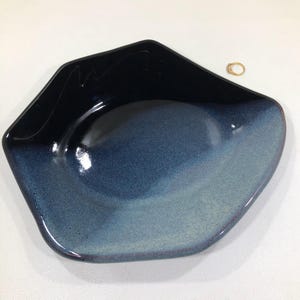May include: A ceramic bowl with a unique, asymmetrical shape. The bowl features a gradient of blue hues, transitioning from a deep navy at the rim to a lighter shade at the center. A small gold ring rests on the white surface next to the bowl.
