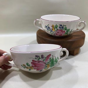 May include: Two white ceramic soup bowls with floral designs. Each bowl has a burgundy rim and two handles. One bowl is on a small wooden stand. The floral patterns include pink roses, blue flowers, and green leaves.